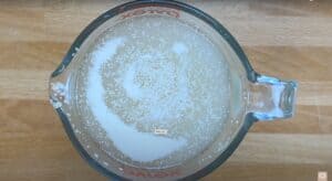 Measuring cup with bubbling yeast mixture on wooden surface, essential for baking bread dough.